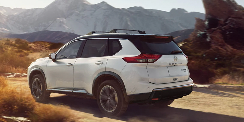 White Nissan Rogue SUV driving on a dirt trail with mountains in the background.