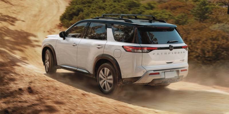 rear left angle view of a white 2024 nissan pathfinder with a black rook driving on a dirt road