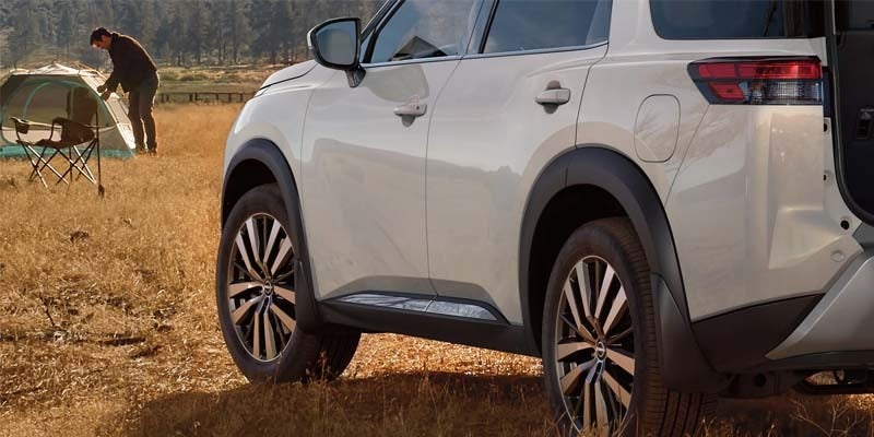 left hand side rear view of a white 2024 nissan pathfinder parked in a field and in the background you can see a man setting up a tent