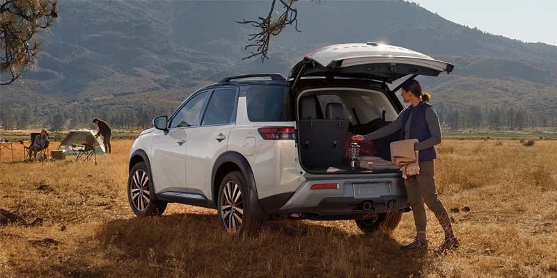 in the foreground there is a white 2024 nissan pathfinder parked in a filed and a woman has the hatchback open and pulling out gear while in the background there is a man setting up a tent