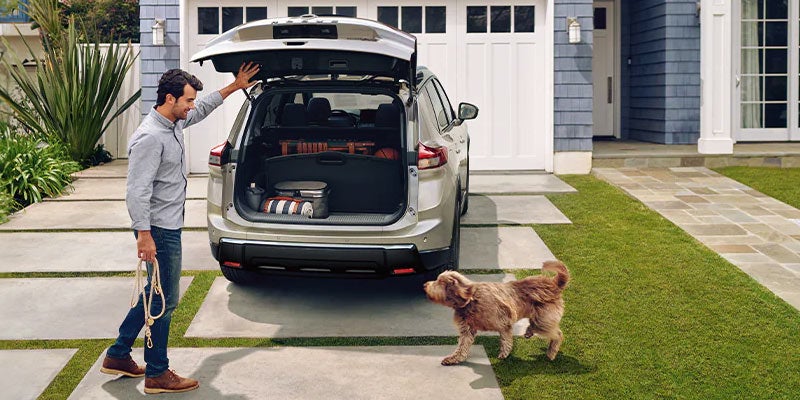 a man signaling his dog to jump in the hatchback trunk of a 2024 nissan rogue that is parked in a driveway