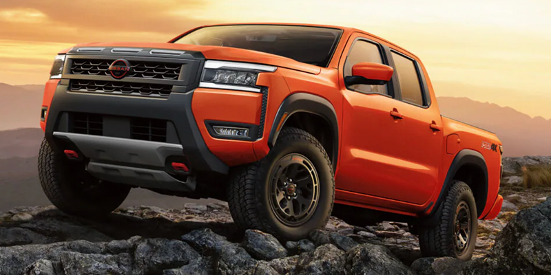 Orange Nissan Frontier pickup truck parked on rocky terrain during a mountain sunset