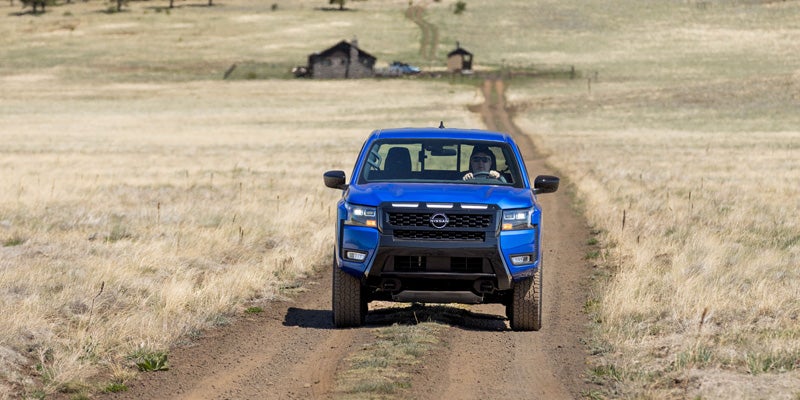 Blue 2027 Nissan Frontier pickup truck driving on a dirt trail through an open grassland landscape